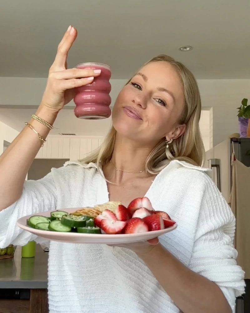 Person holding a pink smoothie in one hand and a plate of sliced strawberries and cucumbers in the other, smiling in a kitchen setting. They are wearing a white top and have long blonde hair.
